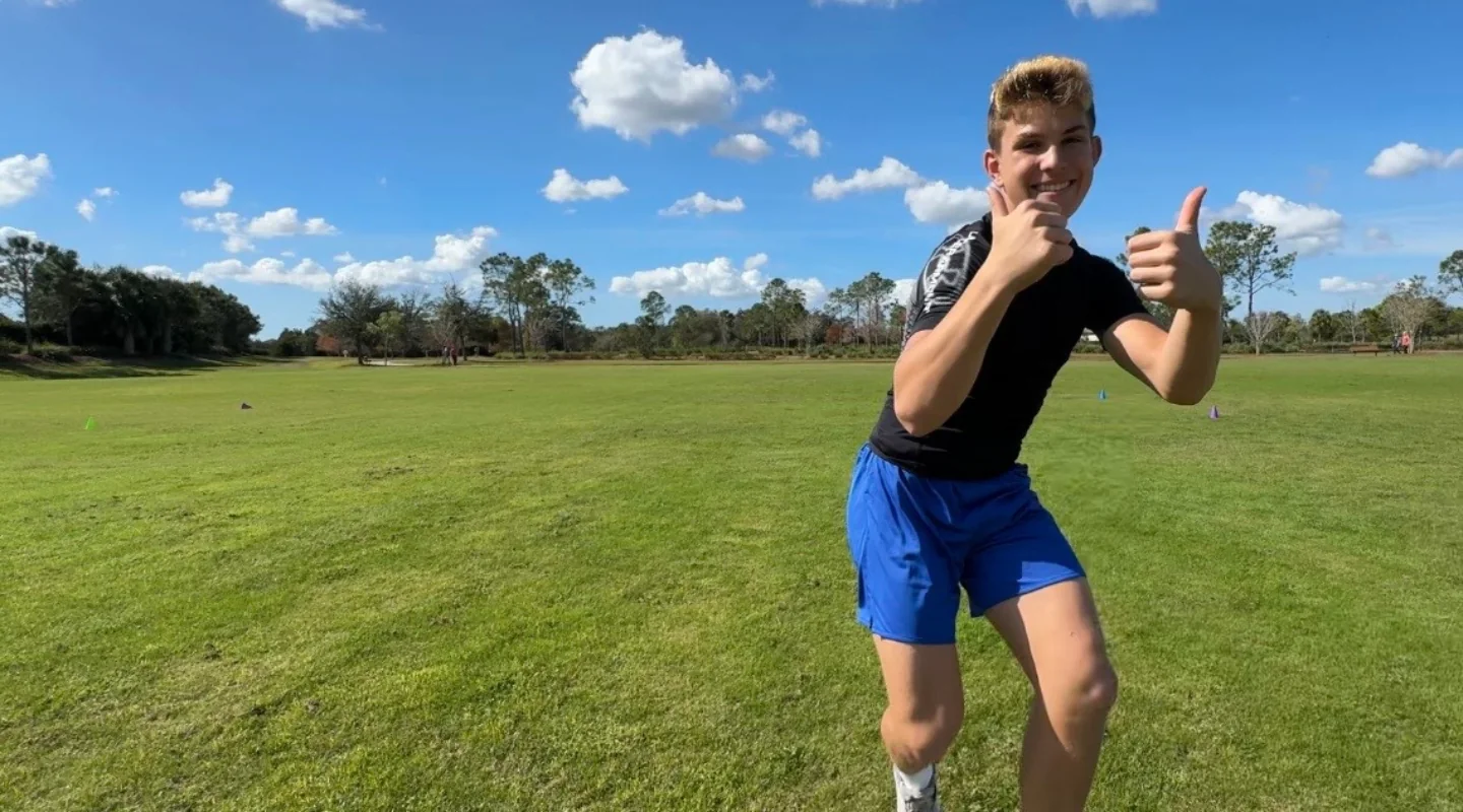 Child smiling and giving two thumbs up in a sunny field.